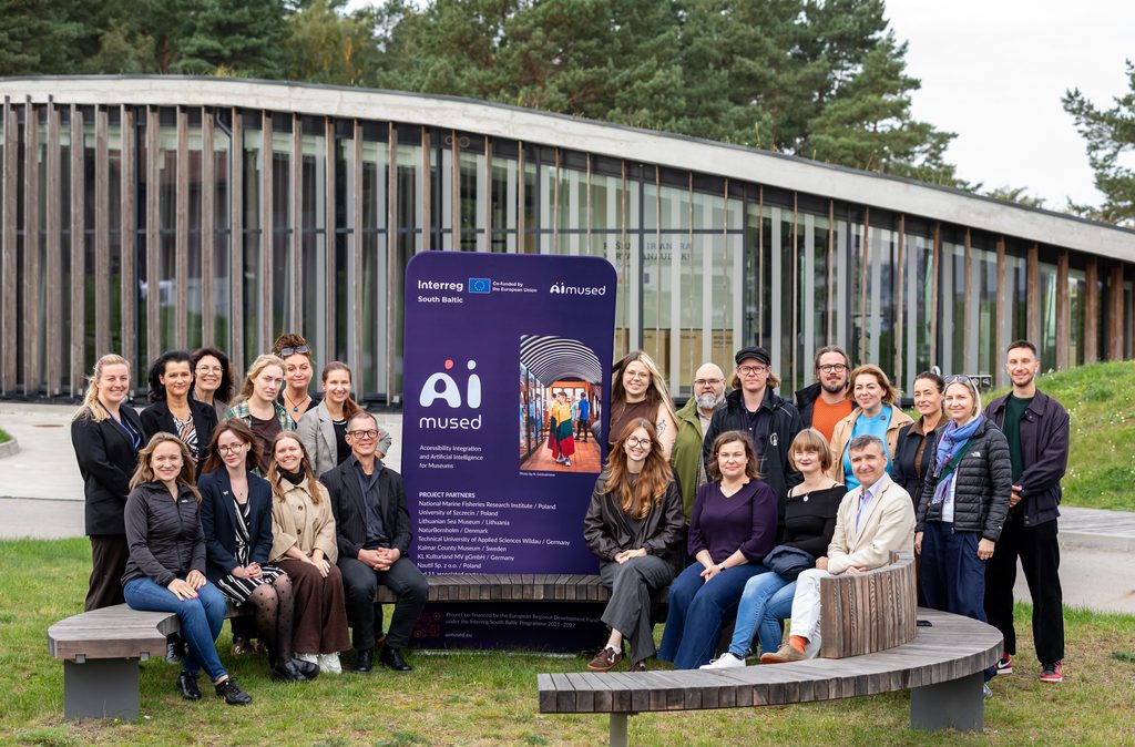 Zdjęcie grupowe partnerów projektu zgromadzonych wokół ścianki tekstylnej z logo projektu i logo programu Interreg South Baltic. 