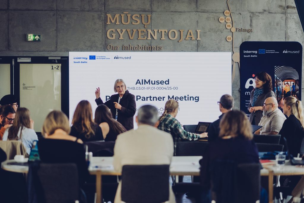 AIMused partner meeting in Klaipėda, with a speaker presenting to project partners seated around tables in front of a large presentation screen.