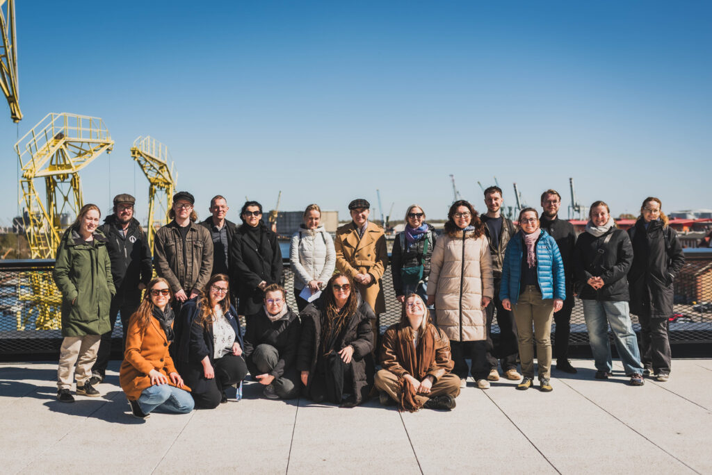 A group of AIMused project partners posing outdoors during a study visit in Szczecin, standing and sitting in rows on a terrace by the waterfront. Behind them are industrial port elements, including yellow cranes and dock structures, under a clear blue sky.