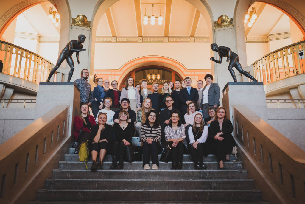 A group of AIMused project partners posing together on a wide staircase inside a museum during a study visit in Szczecin. The participants are arranged in several rows, facing the camera and smiling. The interior features high arches, warm lighting, and two dark sculptures of human figures positioned symmetrically on pedestals above the group.