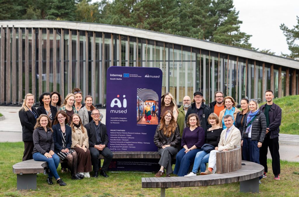 A group of people poses to the photo around a textile board with AIMused project branding.